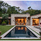Experience “Shaping the World As A Home” by Rizzoli—a showcase of a stone villa with expansive windows, a central outdoor lounge, rectangular pool, and inviting lounge chairs nestled amid lush landscaped greenery and trees. This residence masterfully fuses traditional architecture with contemporary indoor-outdoor living, offering an elegant retreat designed by renowned California architects.