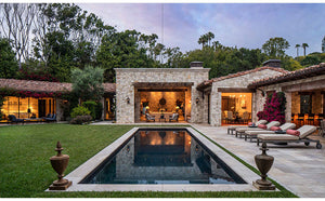 Experience “Shaping the World As A Home” by Rizzoli—a showcase of a stone villa with expansive windows, a central outdoor lounge, rectangular pool, and inviting lounge chairs nestled amid lush landscaped greenery and trees. This residence masterfully fuses traditional architecture with contemporary indoor-outdoor living, offering an elegant retreat designed by renowned California architects.