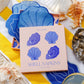 Piecework Shell Cocktail Napkins: a box of four blue shell-shaped linen napkins surrounded by seashells on a yellow and white striped towel.