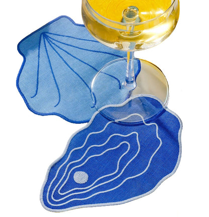 Two blue linen Shell Cocktail Napkins shaped like seashells under a stemmed glass of white wine.