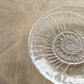 The Shell Serveware Collection by Simon Pearce features a clear shell-shaped glass bowl with a spiral seashell design—ideal as a summer host gift or for beach-inspired decor.