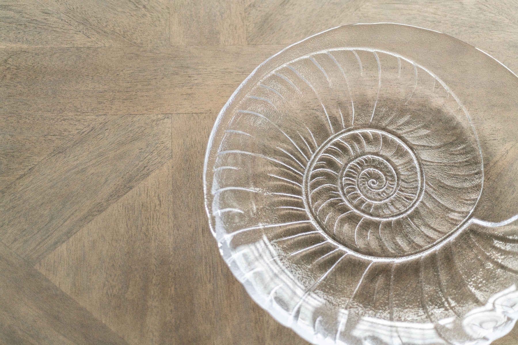 The Shell Serveware Collection by Simon Pearce features a clear shell-shaped glass bowl with a spiral seashell design—ideal as a summer host gift or for beach-inspired decor.