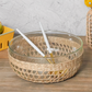A Be Home Removable Seagrass Cage rests on a wooden surface, its glass bowl encased in handwoven rattan and paired with two white and gold utensils, adding rustic charm to any setting.