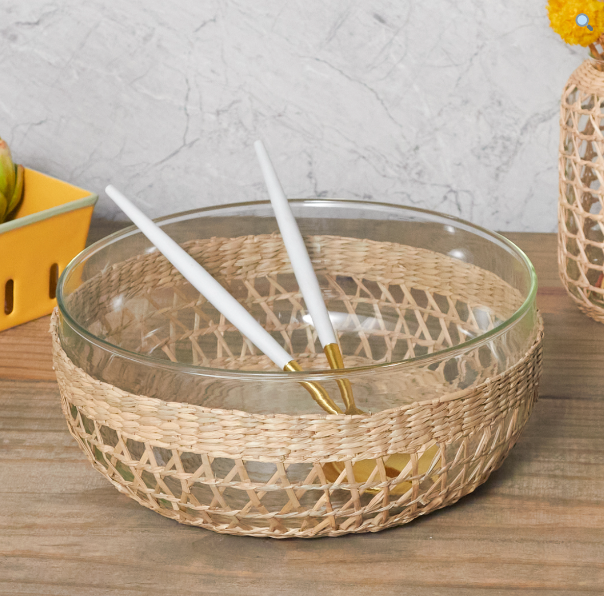 A Be Home Removable Seagrass Cage rests on a wooden surface, its glass bowl encased in handwoven rattan and paired with two white and gold utensils, adding rustic charm to any setting.