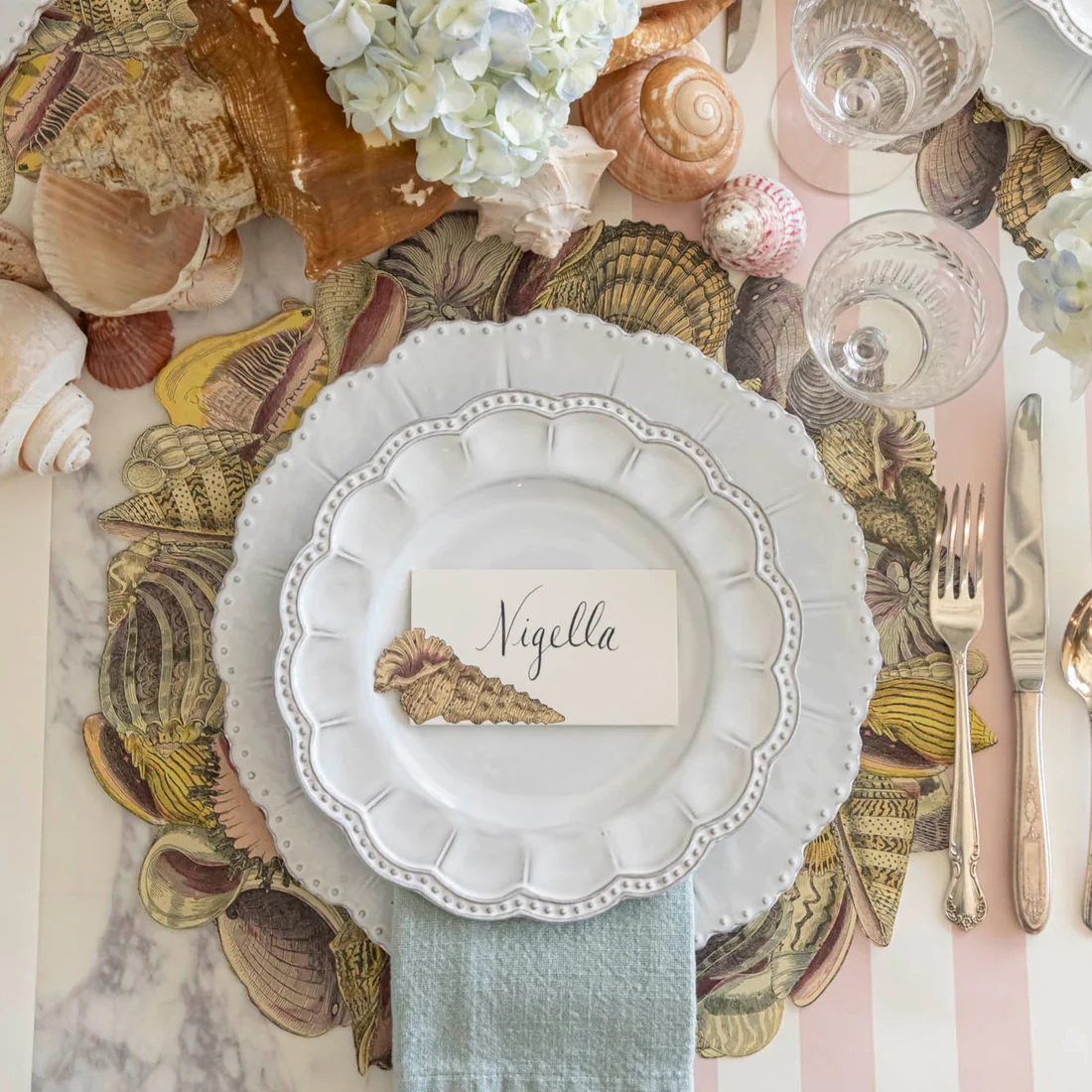 A table set with scalloped white plates, blue napkin, seashell placemat, glassware, and a Hester & Cook Shell Place Card from the Shell Place Card Set labeled "Nigella"—ideal for elegant seaside soirées.