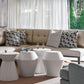 A modern living room features a beige sectional, patterned and solid pillows, three white side tables—including the Hickory Chair Brooke Spot Table—and a gray armchair by large windows overlooking a pool and garden for relaxed elegance.