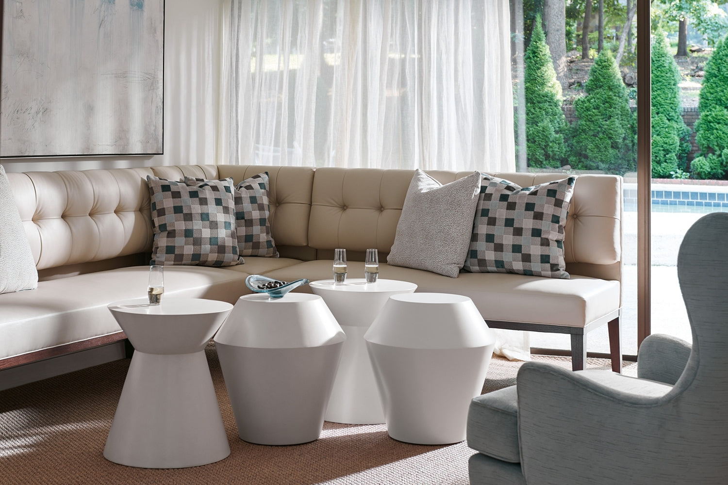 A modern living room features a beige sectional, patterned and solid pillows, three white side tables—including the Hickory Chair Brooke Spot Table—and a gray armchair by large windows overlooking a pool and garden for relaxed elegance.