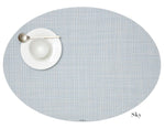 The Chilewich Oval Placemat in light blue brings elegance to any meal, shown with a white plate, small bowl, and metal spoon on the left. Easy to clean with a bleach-based cleanser.