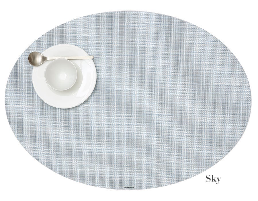 The Chilewich Oval Placemat in light blue brings elegance to any meal, shown with a white plate, small bowl, and metal spoon on the left. Easy to clean with a bleach-based cleanser.