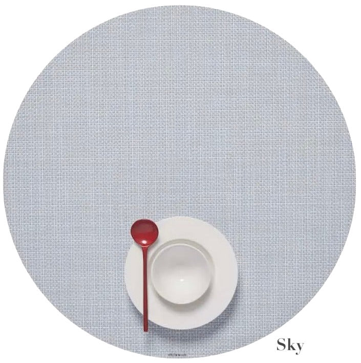 A white cup and saucer with a red spoon sit on a light blue Chilewich Round Placemat, made from phthalate-free Terrastrand yarns, with "Sky" labeled in the bottom right corner.