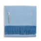 Alicia Adams Alpaca’s Classic Herringbone Throw Collection features a folded light blue baby alpaca blanket with a subtle herringbone pattern, darker blue fringe, and a small fabric label—fair-trade made in Peru.