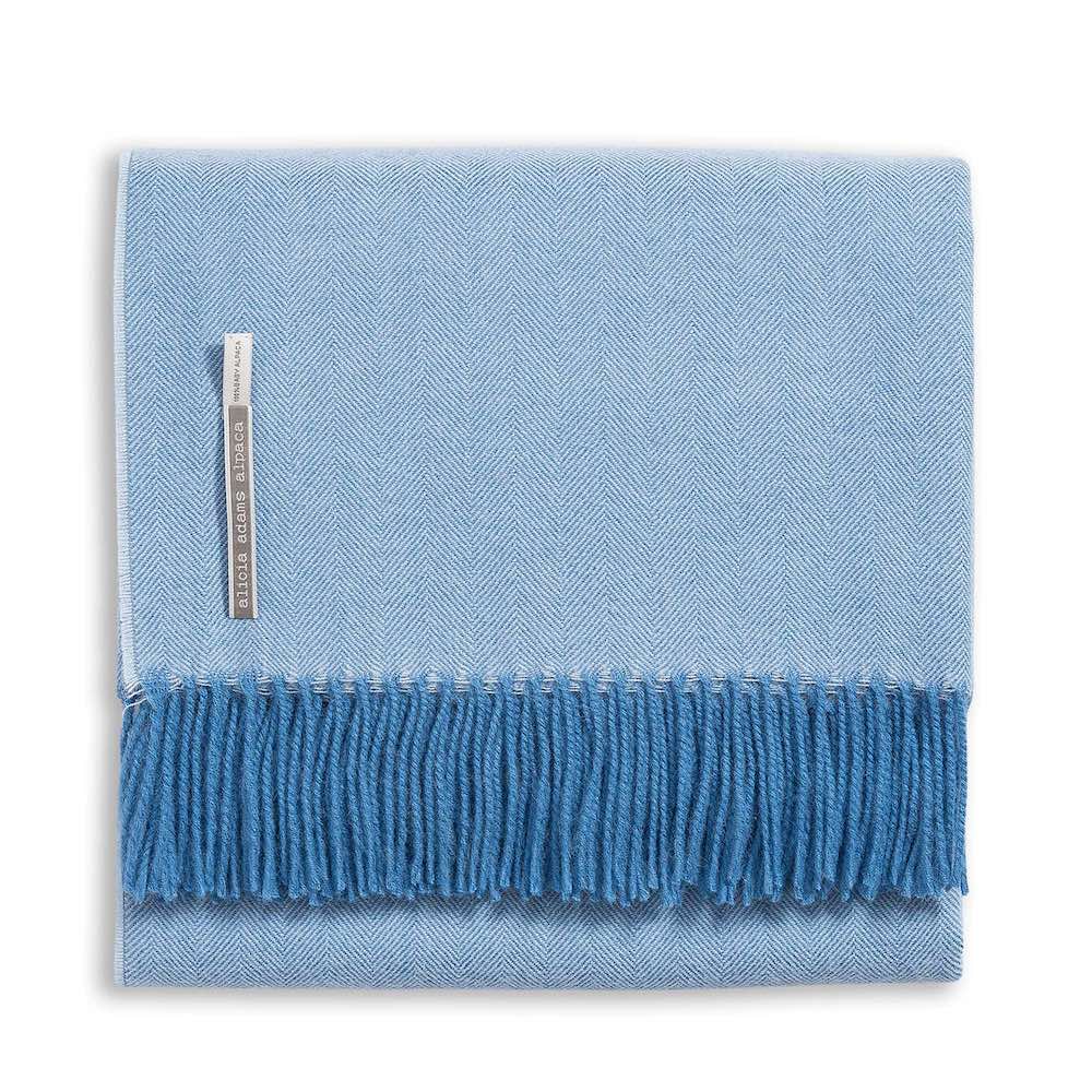 Alicia Adams Alpaca’s Classic Herringbone Throw Collection features a folded light blue baby alpaca blanket with a subtle herringbone pattern, darker blue fringe, and a small fabric label—fair-trade made in Peru.