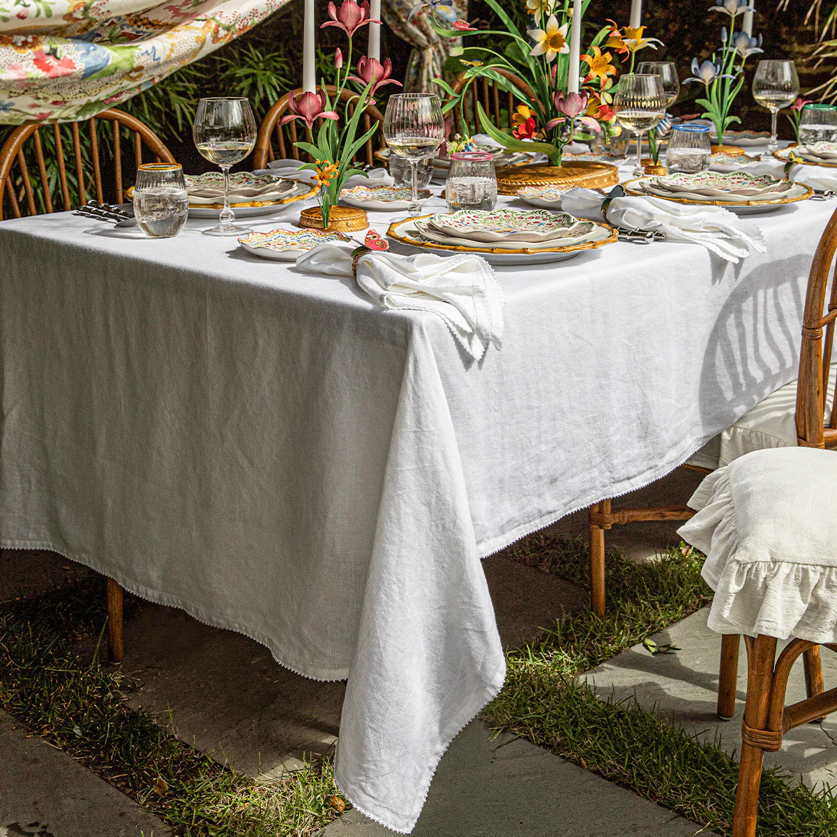A Juliska Berry Scallop Tablecloth dresses a square outdoor dining table, set with plates and glasses. Wooden chairs, floral centerpieces, and surrounding greenery complete the charming, relaxed elegance inspired by Portugal.