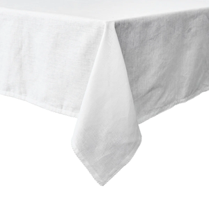 The Juliska Berry Scallop Tablecloth, in white linen and crafted in Portugal, features an elegant scallop trim and drapes gracefully over the corner of a rectangular table against a plain background.