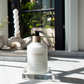 A bottle of hand lotion rests on a Lucite Soap & Candle Tray Collection tray by L’Avant Collective atop a marble countertop.