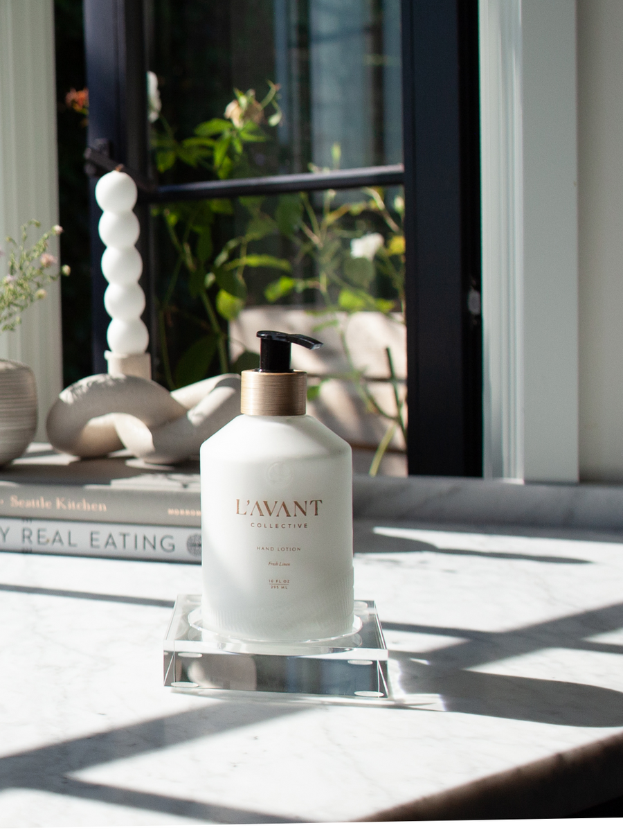 A bottle of hand lotion rests on a Lucite Soap & Candle Tray Collection tray by L’Avant Collective atop a marble countertop.