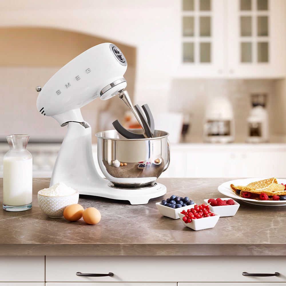 A white SMEG Stand Mixer from the Smeg Collection with a die-cast aluminum body sits on the kitchen counter surrounded by flour, eggs, milk, berries, and pancakes. Its 10 variable speeds and planetary action ensure perfect mixing every time.