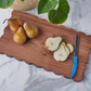 Three whole pears and pear slices with visible seeds rest on the etúHOME Scalloped Serving Board, Medium Rectangle, beside a blue-handled knife; green leaves are partly visible above.