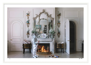 A person in a spacesuit stands next to a fireplace in an elegantly decorated room with an ornate mirror and vases, reminiscent of a Palm Beach estate. Two doors are slightly open on either side of the fireplace, and framed white wood accents add a touch of refinement to the setting. They are holding Nick Mele Fine Art S'mores by Nick Mele Photography.