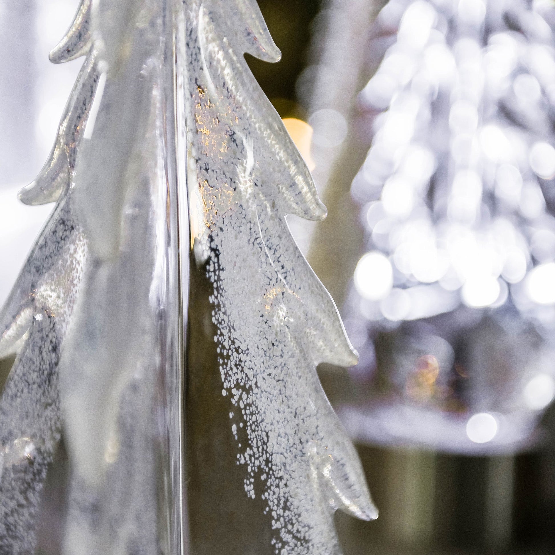 Close-up of a frosted glass pine tree ornament from Simon Pearce’s Snowy Branches Evergreen Tree Collection, with blurred lights in the background evoking Vermont’s winter woods.