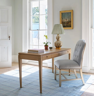 The Sofia Desk in oak by Bunny Williams sits by tall windows, styled with books, a plant, and a decorative lamp. A tufted chair rests at the desk atop a light blue rug, creating a bright and inviting workspace.