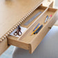 An open drawer of the Bunny Williams Sofia Desk in oak holds scissors, a ruler, markers, pens, pencils, and white paper on its light-colored surface.