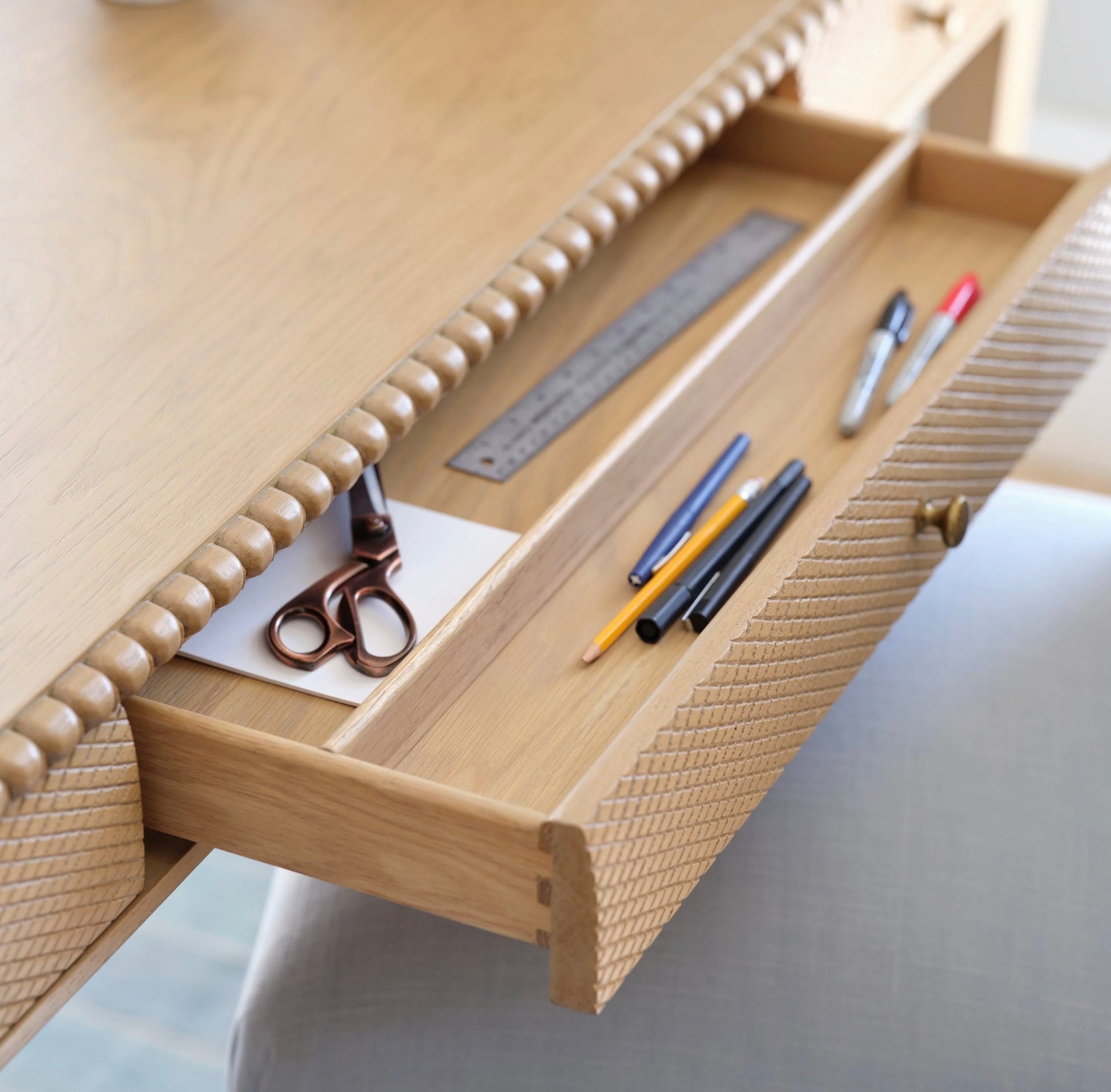 An open drawer of the Bunny Williams Sofia Desk in oak holds scissors, a ruler, markers, pens, pencils, and white paper on its light-colored surface.