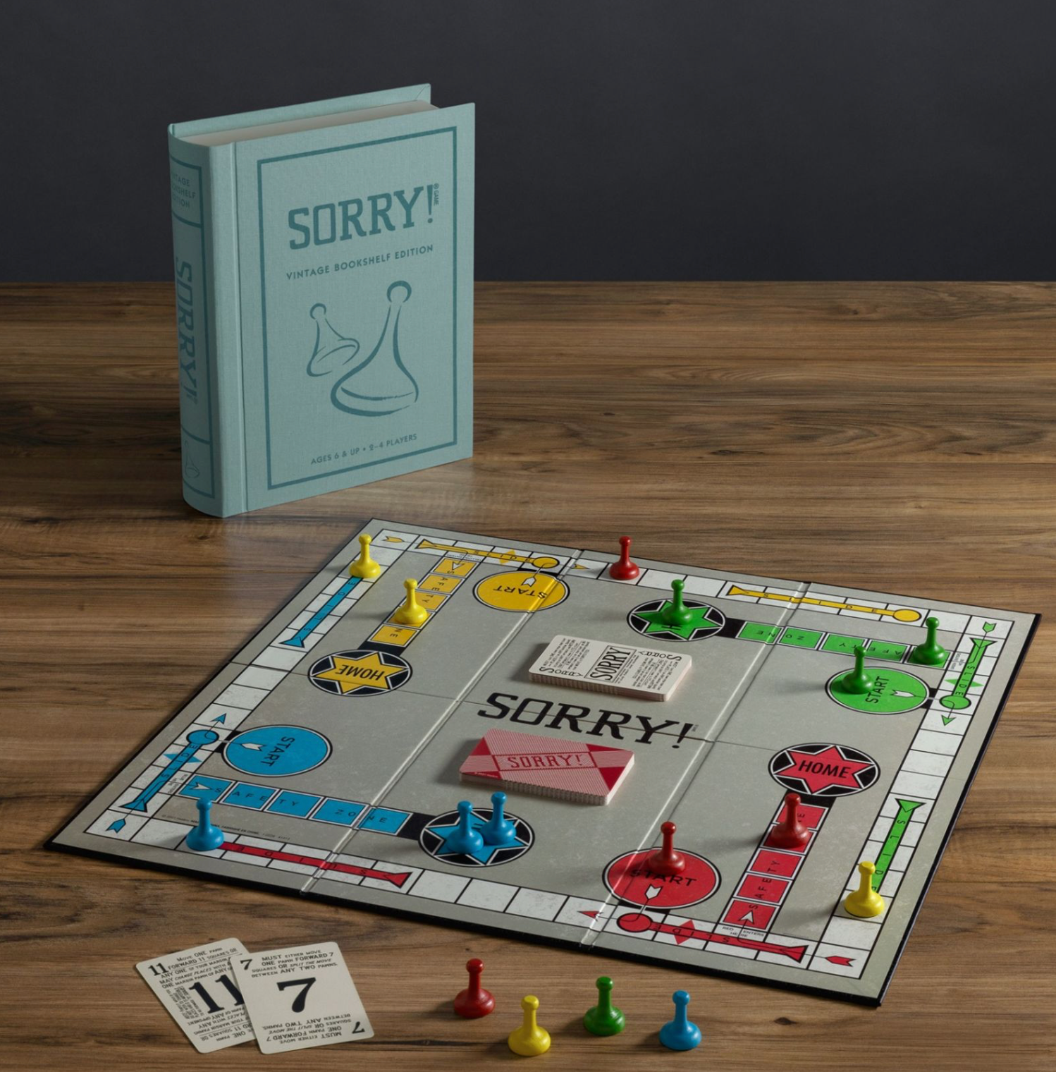 A collectible WS Game Company Sorry! Vintage Bookshelf Edition is placed on a wooden table, showcasing the classic game board with pieces, cards, and a rule book elegantly styled as a vintage book standing upright in the background.