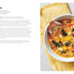 A bowl of chunky vegetable soup—brimming with kale, beans, tomatoes, and carrots—is served with bread on a yellow napkin beside a printed recipe page from The Soup Book by Penguin Random House, showcasing nourishing recipes perfect for any season.