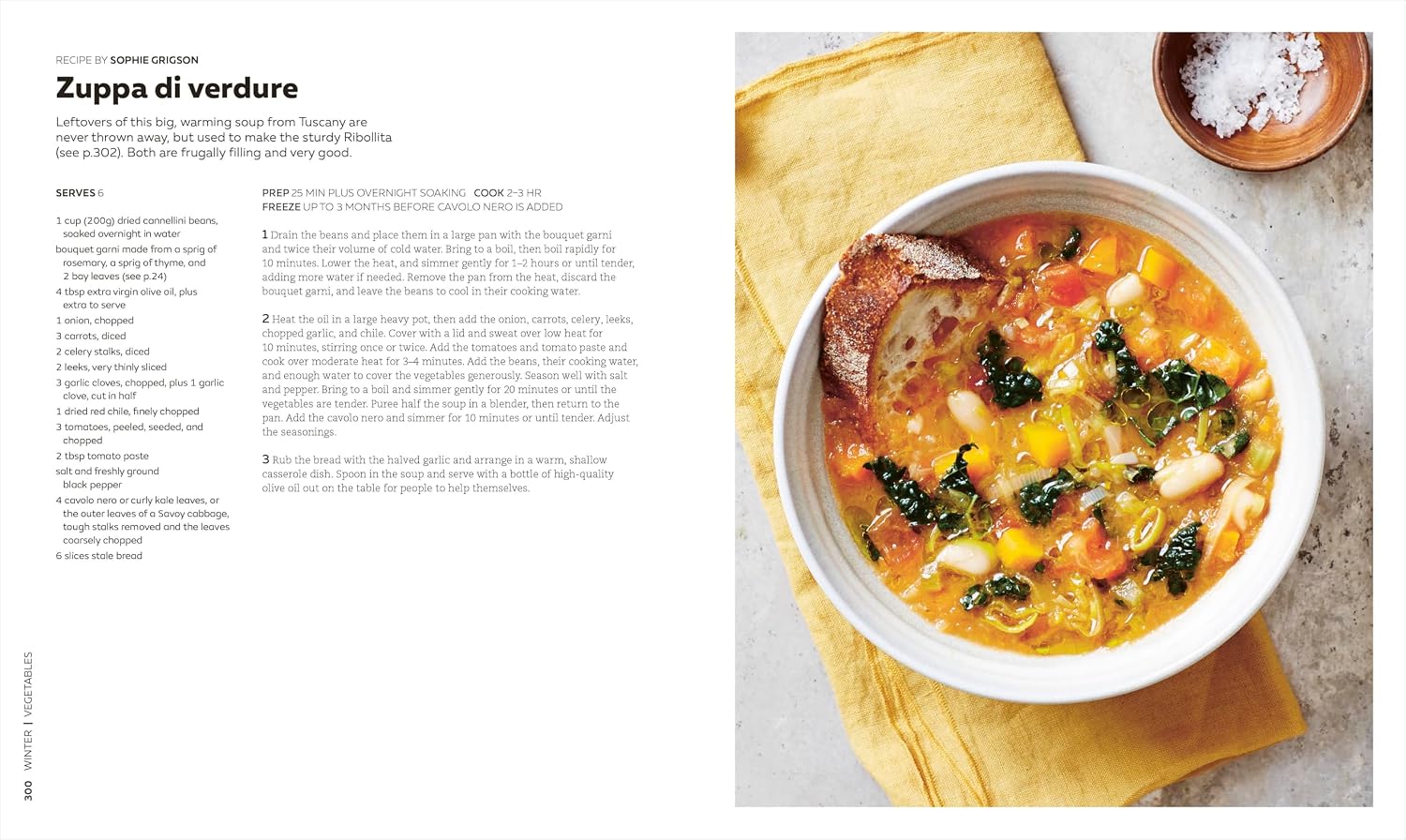 A bowl of chunky vegetable soup—brimming with kale, beans, tomatoes, and carrots—is served with bread on a yellow napkin beside a printed recipe page from The Soup Book by Penguin Random House, showcasing nourishing recipes perfect for any season.