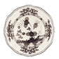 The Oriente Italiano Albus Dinnerware Collection by Ginori 1735 features ceramic plates with scalloped edges and intricate black and white floral and landscape designs, inspired by Ginori 1735 craftsmanship.