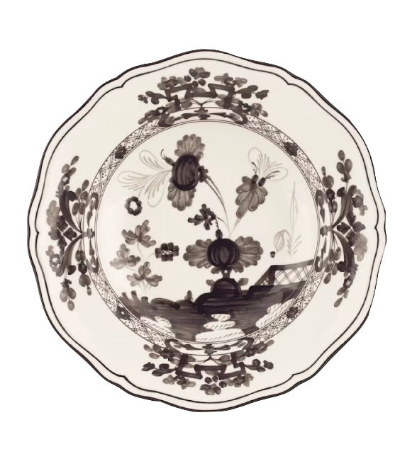 The Oriente Italiano Albus Dinnerware Collection by Ginori 1735 features ceramic plates with scalloped edges and intricate black and white floral and landscape designs, inspired by Ginori 1735 craftsmanship.