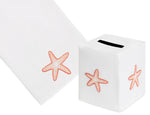 The Haute Home Playful Starfish Bath Collection in Coral features a white tissue box cover and towel, both crafted from Italian linen and adorned with hand embroidered orange starfish designs.