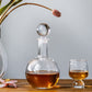 The Benson Drinkware Collection by Simon Pearce features a round glass decanter with amber liquid beside a matching glass on wood, its clean lines creating a geometric statement next to a flower vase in the background.