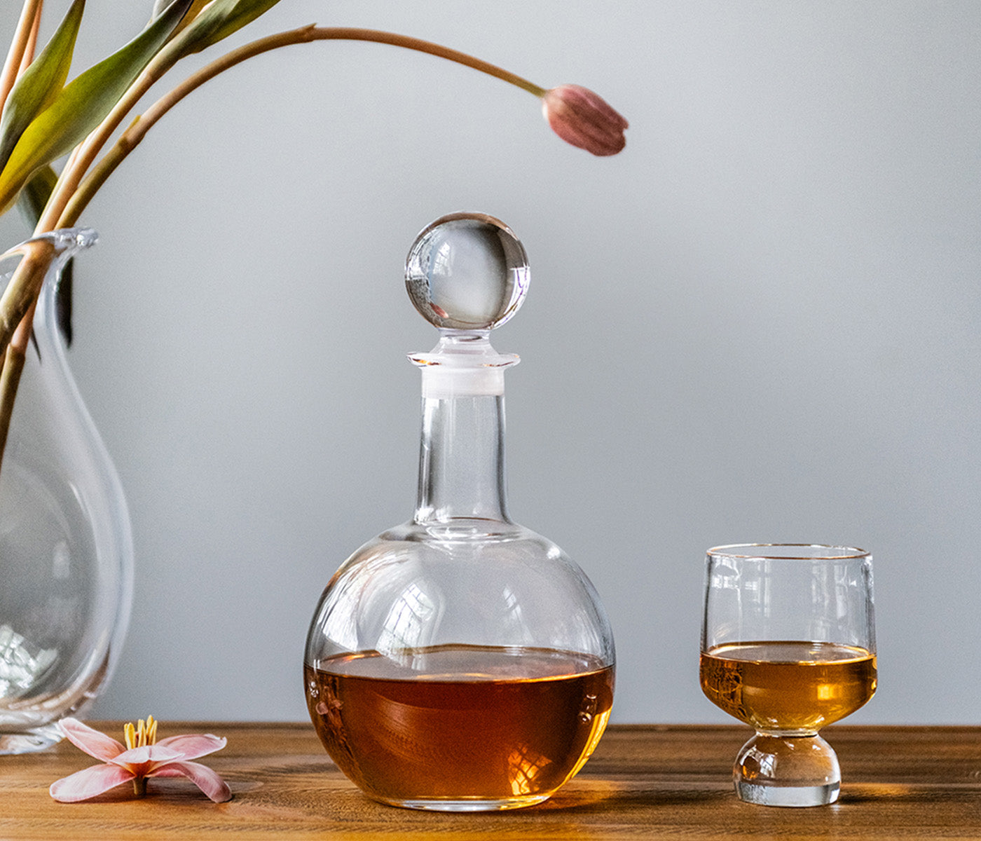 The Benson Drinkware Collection by Simon Pearce features a round glass decanter with amber liquid beside a matching glass on wood, its clean lines creating a geometric statement next to a flower vase in the background.
