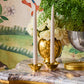 The Olivia Brass Taper Candleholder Set by Aerin, holding two white tapers, is displayed on marble with a vase of white roses, a silver planter with greenery, and a gold container, set against floral wallpaper for an Art Deco touch.