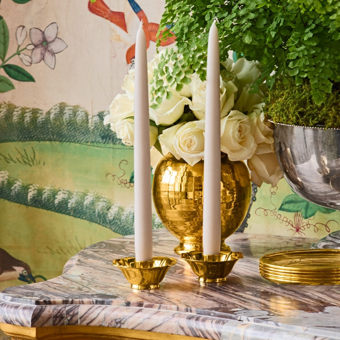 The Olivia Brass Taper Candleholder Set by Aerin, holding two white tapers, is displayed on marble with a vase of white roses, a silver planter with greenery, and a gold container, set against floral wallpaper for an Art Deco touch.