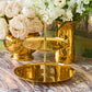 The Sintra 2-Tier Brass Stand by Aerin is shown on a marble surface next to gold vases with white flowers, making it an elegant choice for stylish cake service.
