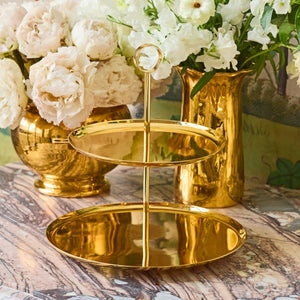 The Sintra 2-Tier Brass Stand by Aerin is shown on a marble surface next to gold vases with white flowers, making it an elegant choice for stylish cake service.