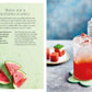 A glass of pink Spritz Cocktails by Simon & Schuster, served over ice with a touch of Italian amaro, rests on a green coaster beside a plate of fresh watermelon slices. In the background, a candle lantern casts a warm glow.