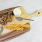 The Be Home Acacia Wood Rectangular Board with Handle Collection displays cheese, a cheese knife, fruit and nut bread slices, and two twisted breadsticks on a light surface.