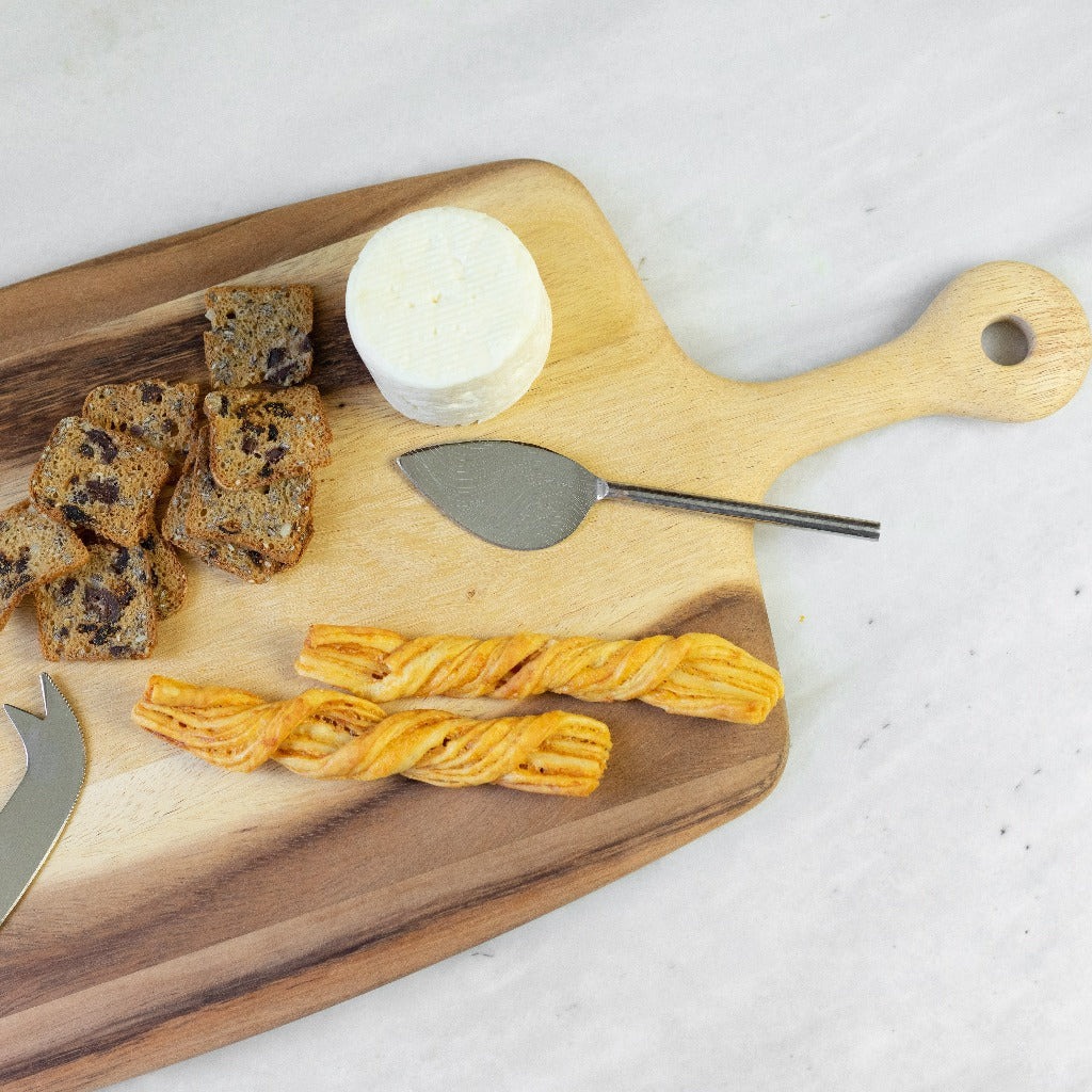 The Be Home Acacia Wood Rectangular Board with Handle Collection displays cheese, a cheese knife, fruit and nut bread slices, and two twisted breadsticks on a light surface.
