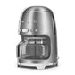 The Smeg Drip Filter Coffee Machine Collection features a silver finish, glass carafe, digital display with buttons, keep-warm function, and a side water level indicator.