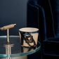 A Baobab Collection Stones Lazuli Candle by Baobab with a dark marbled container sits on a glass side table with a metallic base, next to a navy blue velvet chair.