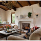 Living room with brown sofas, a lit fireplace, coffee tables adorned with books and flowers—including *Shaping the World As A Home* by Rizzoli—large windows, and exposed wood beam ceiling exemplify beautiful traditional architecture. Candles and a potted plant decorate the mantel.