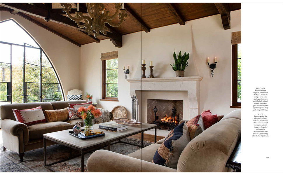 Living room with brown sofas, a lit fireplace, coffee tables adorned with books and flowers—including *Shaping the World As A Home* by Rizzoli—large windows, and exposed wood beam ceiling exemplify beautiful traditional architecture. Candles and a potted plant decorate the mantel.