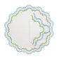 A round white linen placemat and folded napkin from the Sugus Table Linen Collection, featuring blue scalloped edges and green, pink, and yellow decorative embroidery.