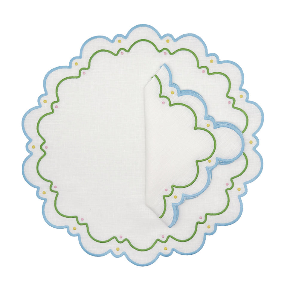 A round white linen placemat and folded napkin from the Sugus Table Linen Collection, featuring blue scalloped edges and green, pink, and yellow decorative embroidery.