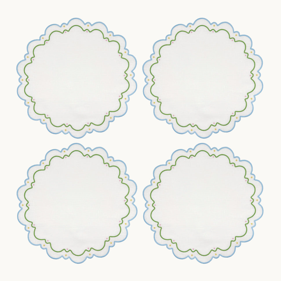 Four round white embroidered Sugus Table Linen Collection placemats with scalloped edges and blue and green decorative detailing.