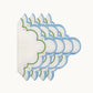 A stack of Sugus Table Linen Collection napkins, featuring white linen with scalloped edges embroidered in blue, green, and small yellow and pink dots.